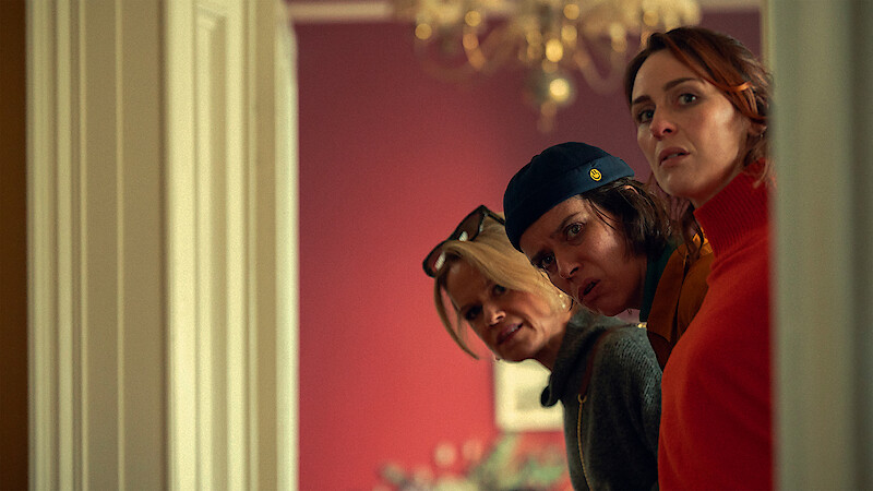 Three women stand at a hotel reception desk in a warmly lit room, looking serious or suspicious. A newspaper, a bell, and a lamp are on the desk, creating a tense, mysterious atmosphere.