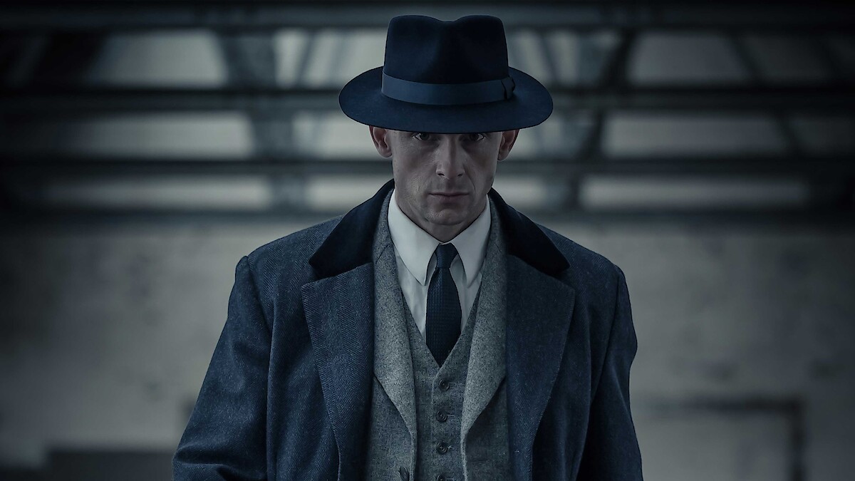 Man in vintage suit and fedora hat standing indoors in a dimly lit, industrial-style room, facing forward with a serious expression. The setting has concrete walls and visible metal beams in the background.