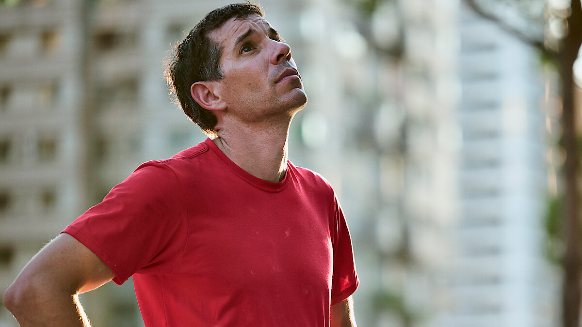 Alex Honnold in a red shirt stands outdoors in an urban setting, looking upwards with a thoughtful expression. Blurred tall buildings and trees are in the background, suggesting a city environment.