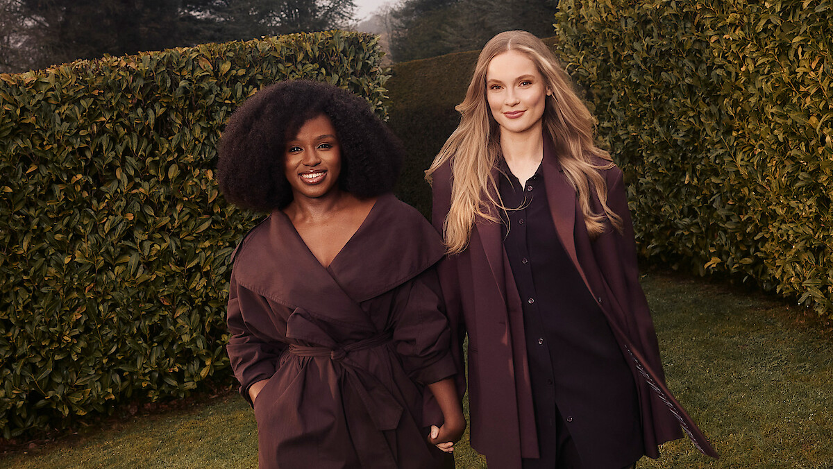 Two women standing outdoors in a garden with tall manicured hedges, wearing stylish, coordinated dark outfits, smiling and holding hands, with soft natural lighting and trees in the background.