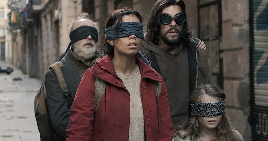 Three adults and a child walk through the street wearing blindfolds.