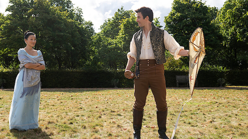 A woman in a light blue dress and a man in period costume with a kite stand on a grassy field in a garden, surrounded by green trees under a partly cloudy sky.