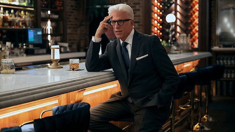 Older man in a dark suit sits at an elegant bar with modern lighting and upscale decor, looking thoughtful and composed.