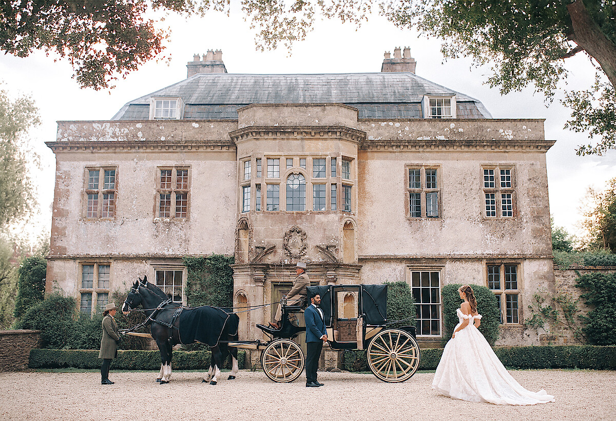 Bride in a white gown stands near a vintage horse-drawn carriage outside an elegant, historic mansion. Lush greenery frames the scene, evoking a romantic, fairytale atmosphere. The mood is dreamy and regal.
