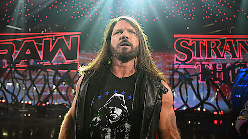 AJ Styles stands in a dramatic wrestling arena, wearing a black vest and T-shirt, with bright red RAW and STRANGER THINGS signs and stage lights in the background.