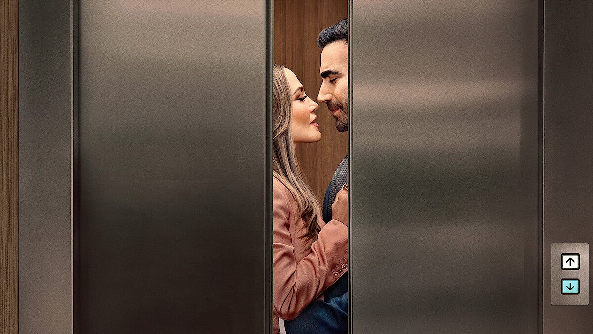 A couple standing close together inside an elevator, gazing at each other affectionately as the stainless steel elevator doors partially close, suggesting a romantic or intimate moment in a modern building setting.