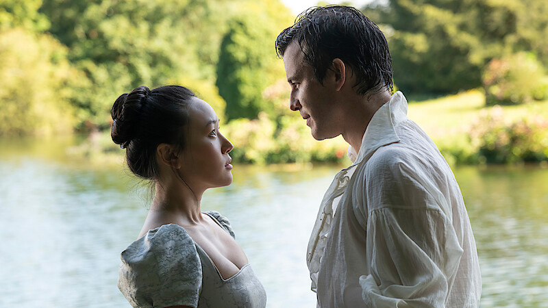 A man and woman in period clothing stand closely facing each other near a lake, surrounded by greenery and trees on a bright day, suggesting a romantic or dramatic moment outdoors.