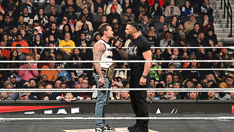 Two wrestlers face off in a packed arena wrestling ring, one holding a championship belt and the other speaking into a microphone as a large audience watches the dramatic confrontation.