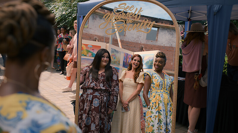 Three women smiling and posing together in front of a decorative mirror at an outdoor event, with other people and a tent in the background. The mirror has the words "See Yourself in Serenity" written on it.