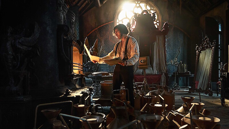 A man studies papers in a dark, ornate room filled with scientific equipment and warm, dramatic light streaming through a stained-glass window, creating a mysterious and intense mood.