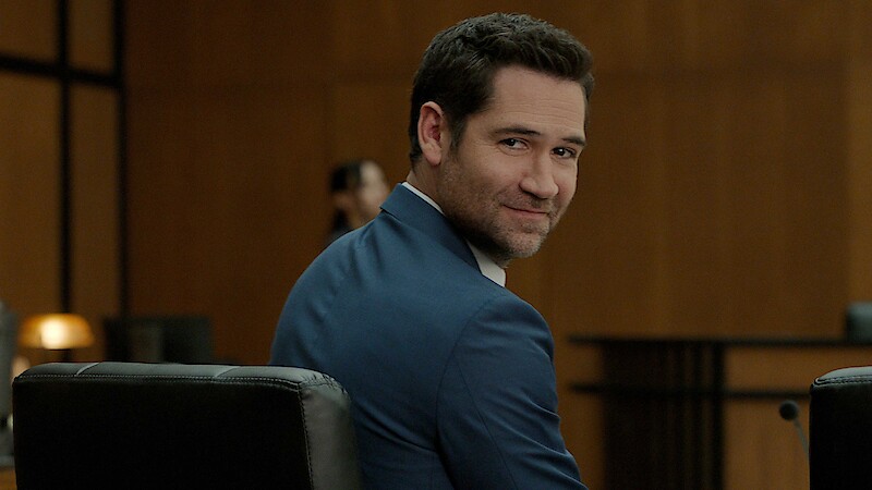 Man in a blue suit sitting in a modern courtroom setting, turning to smile at the camera with wood-paneled walls and blurred figures in the background.
