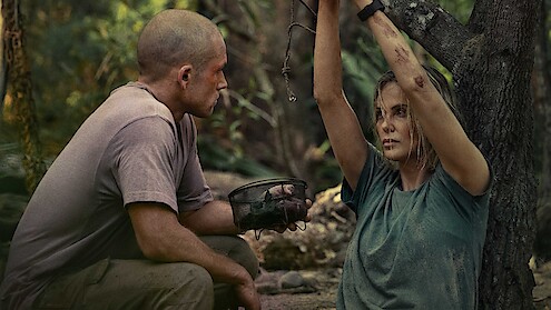 A man kneels beside a woman tied to a tree in a dense forest, holding a canteen. Both look dirty and tense, suggesting a survival or hostage situation in a rugged, outdoor setting.