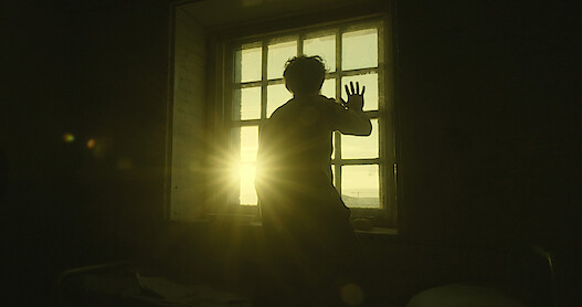   A man puts his hand to a window as the sun rises in the distance.