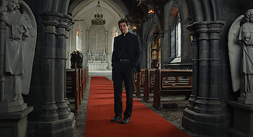 A priest stands on a red carpet in the middle aisle of a dimly lit stone church, flanked by statues, with pews on either side and an altar in the background, creating a solemn and contemplative mood.
