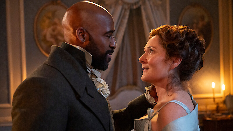 A man and woman in historical clothing gaze at each other in an elegantly decorated, candlelit room with ornate walls and portraits, suggesting a romantic or emotional moment in a period drama setting.