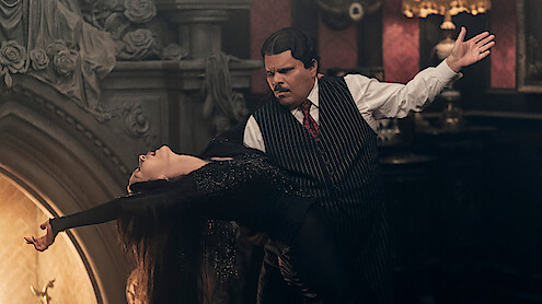 Catherine Zeta-Jones as Morticia Addams and Luis Guzmán as Gomez Addams dancing together in front of a fireplace in 'Wednesday' Season 2.