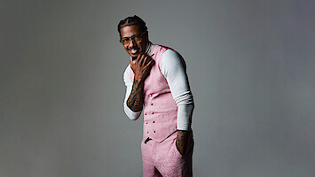 Nick Cannon in pink vest and pants over white turtleneck poses confidently in a studio with a neutral gray background, smiling while touching his chin, wearing glasses, and showing tattooed arms.