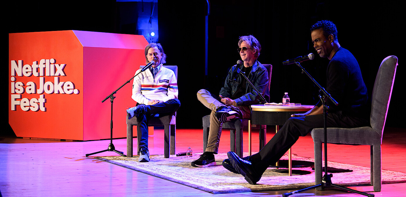 David Spade, Dana Carvey and Chris Rock sit on stage with microphones at Netflix is a Joke Fest, with a red event sign, round table, water bottles, and casual conversation in a well-lit theater setting.
