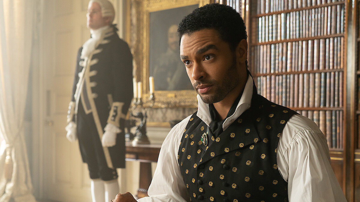 A man in Regency-era clothing sits in a richly decorated library with bookshelves and paintings, while a servant stands in the background near a window and candle holder.