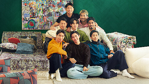 The cast of 'The Boyfriend' Season 2 sit closely together on a colorful sofa in a cozy living room with eclectic pillows, patterned furniture, and a vibrant art piece on a green wall, smiling and posing for the photo.