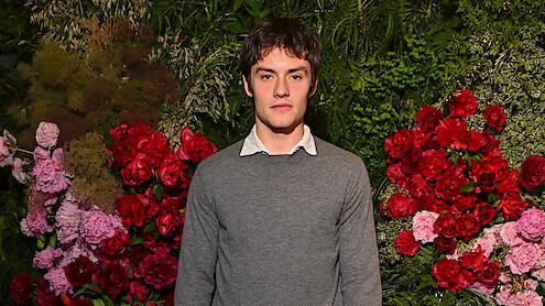 Young man in a gray sweater stands in front of a vibrant floral wall with red and pink flowers and green foliage, creating a natural, garden-like backdrop. Indoor setting, formal or event atmosphere.