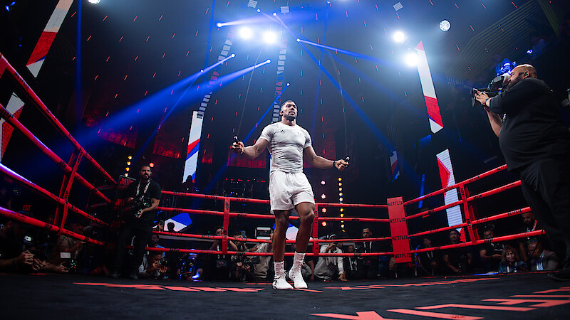 Boxer in a ring celebrates with a raised, gloved fist under dramatic arena lighting, surrounded by a cheering crowd, capturing a victorious and energetic mood.