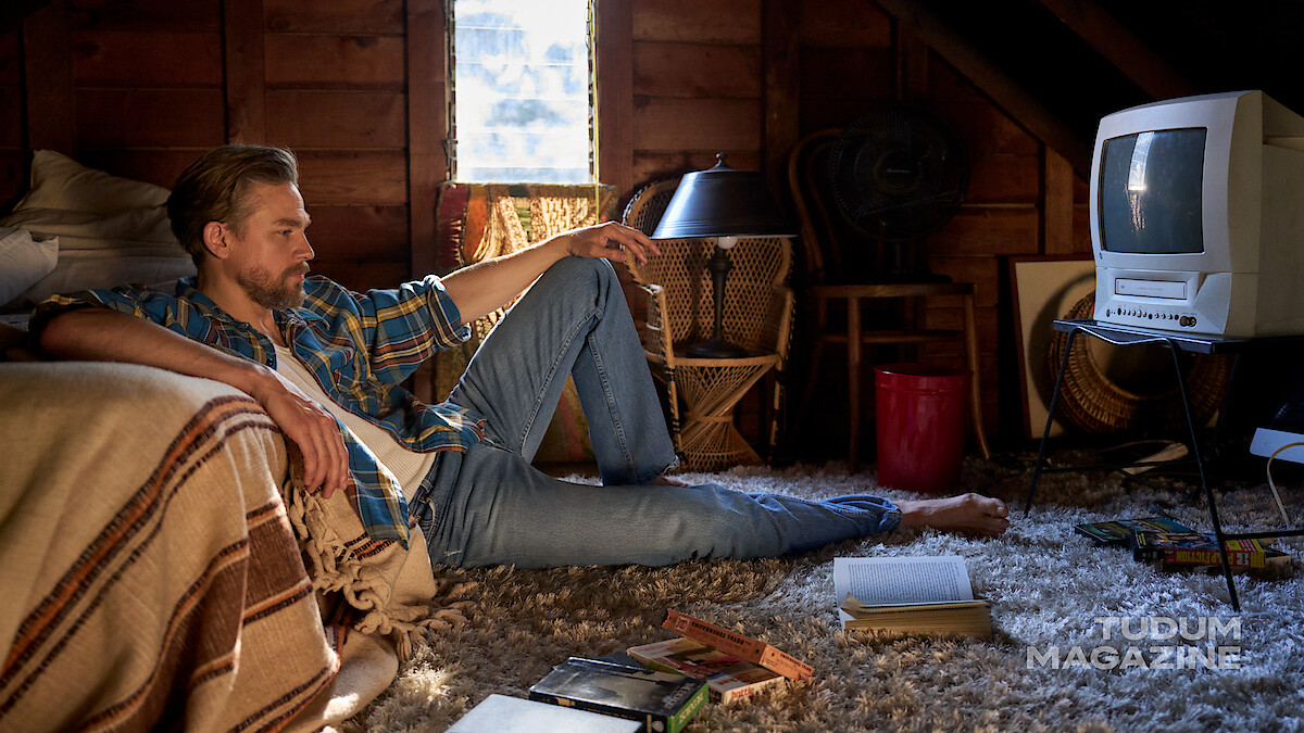 Charlie Hunnam sits in a cabin bedroom on the floor surrounded by VHSs and books, staring into the distance with a contemplative look on his face. 