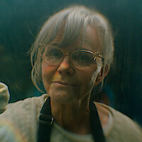 Elderly woman with gray hair and glasses wearing an apron, softly smiling indoors with a blurred background, seen through a slightly foggy or reflective surface.