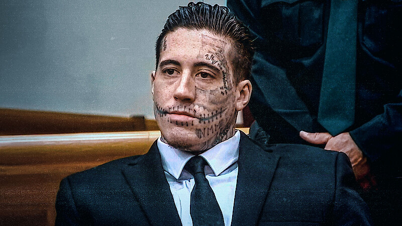 Man with extensive facial tattoos wearing a suit, seated in a courtroom setting with wooden benches and another person in uniform visible in the background.