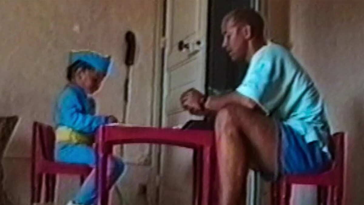 A child in a blue costume sits at a small red table with an adult in casual clothes, inside a simple room with beige walls and a white door, creating a warm and intimate indoor setting.