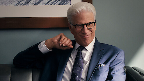 Smiling man with white hair and glasses in a blue suit sits on a leather couch in an office, with a framed picture and blue wall in the background. The atmosphere is professional and relaxed.