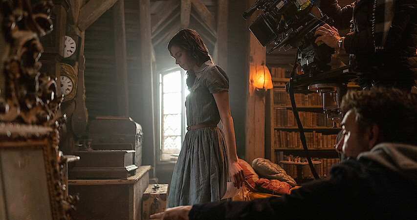 A behind the scenes still of Aria Mia Loberti standing in a room while a camera films the scene