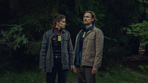 Two people wearing badges and jackets stand side by side in a dense, green forest, holding hands and looking in opposite directions, suggesting a serious or investigative scenario in a natural outdoor setting.