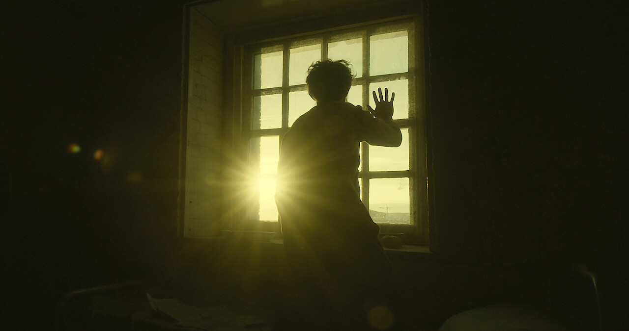   A man puts his hand to a window as the sun rises in the distance.