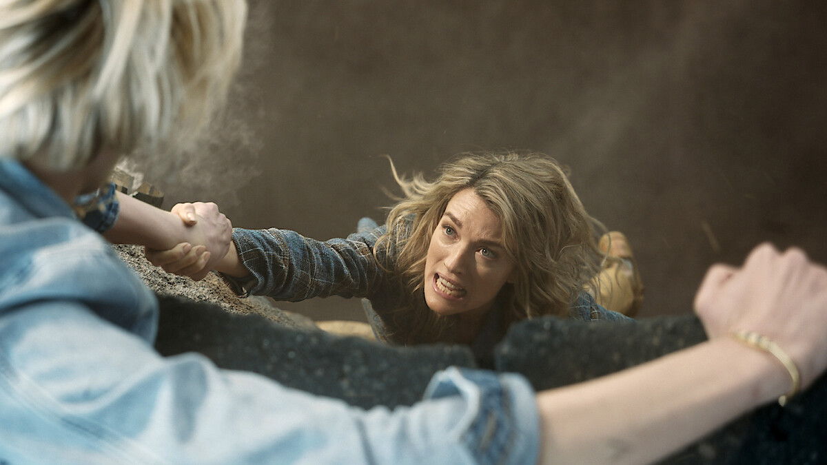 A woman hanging from a ledge, gripping another person's arm, with a distressed expression, surrounded by dust and debris in a tense, dramatic rescue scene.