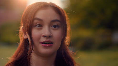 A young woman with long brown hair looks surprised outdoors in a park or garden setting during sunset, with soft natural lighting and a blurred green background.