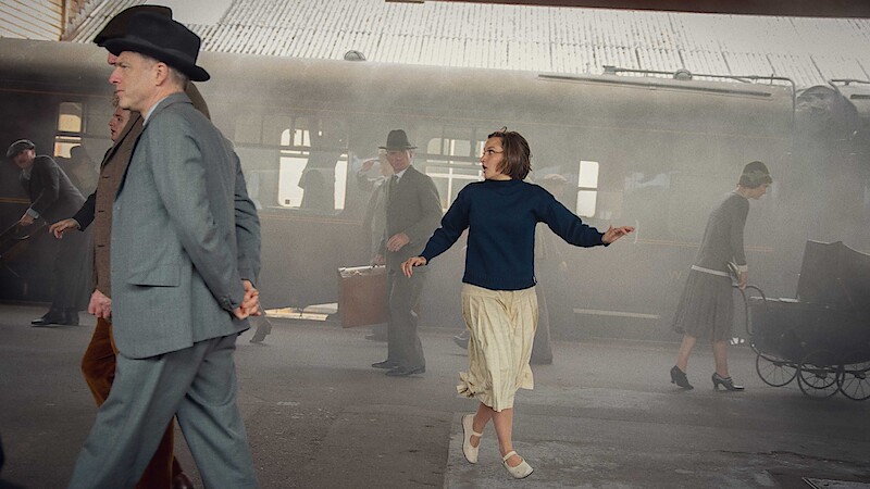 A woman in a skirt and sweater looks around anxiously at a vintage train station as people in old-fashioned clothing walk by, with steam and luggage visible in the background.