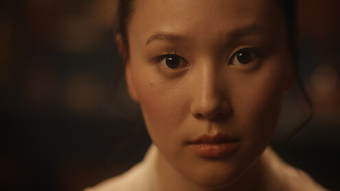 Young woman with neutral expression looking at camera, softly lit face, blurred dark indoor background, close-up portrait.