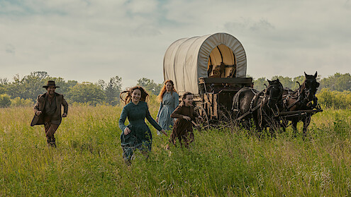 A family runs through a field with a wagon behind them. 