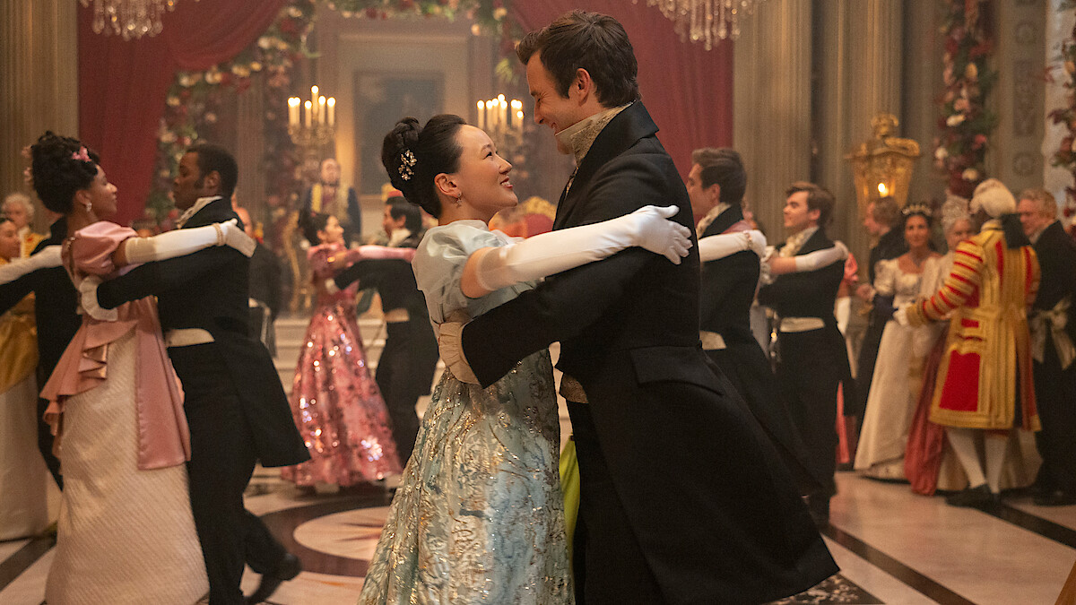A couple in elegant period costumes dance together in a grand ballroom, surrounded by other couples waltzing and elaborately dressed guests, ornate decorations, flowers, and chandeliers creating a festive, historical atmosphere.