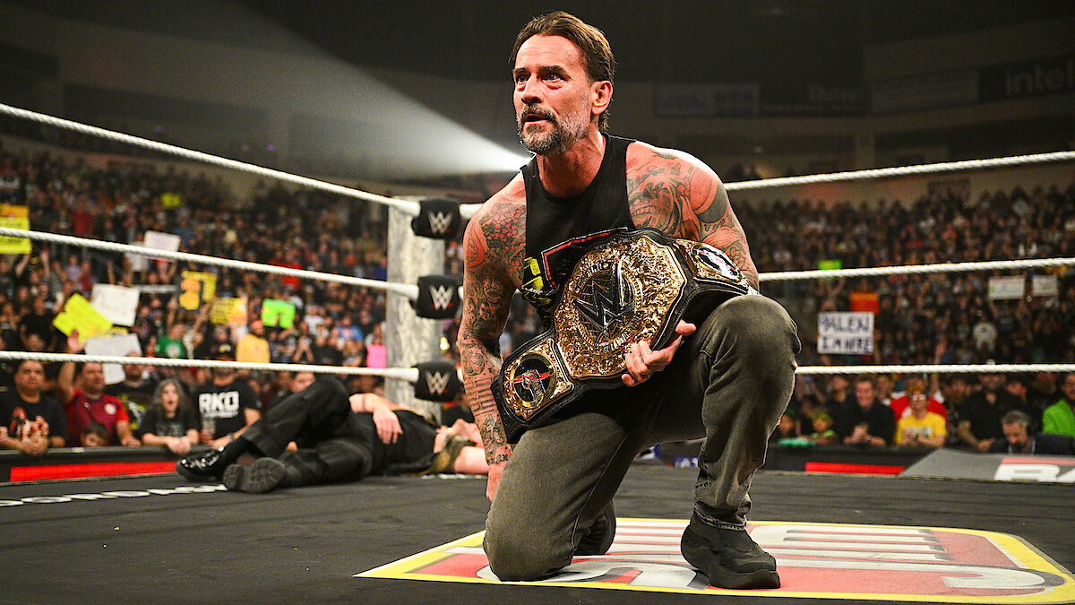 CM Punk kneels in a wrestling ring holding a championship belt, with a cheering crowd and colorful signs in the background during a live event.