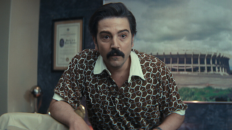 Man with dark hair and mustache in patterned shirt leans forward in an office with framed certificate on wall and large stadium photo in the background.