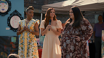 Three women in summer dresses stand together outdoors at an art fair or festival, one holding a microphone. Colorful paintings and a canopy tent are visible in the background.