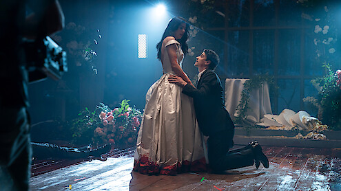 A man in a suit kneels before a woman in a white dress with red trim in a dimly lit, flower-filled room, dramatic lighting highlighting the emotional scene, with petals and decorations scattered on the wooden floor.