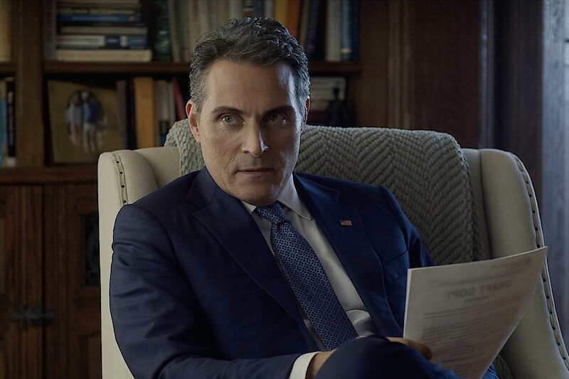 Man in a blue suit sits in an armchair holding a document, in a library or office with bookshelves and framed photo behind him, appearing serious and contemplative.
