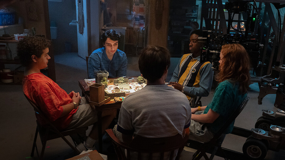 Five teenagers sitting around a table playing a board game in a dimly lit basement, surrounded by various objects and filming equipment, creating a cozy and nostalgic atmosphere.