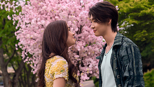 Two young people stand face to face, smiling at each other under a blooming pink cherry blossom tree in a park setting with green foliage in the background.
