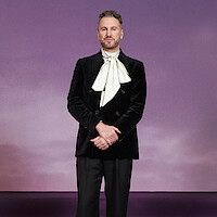 A man stands against a purple gradient background, wearing a black velvet double-breasted jacket with a large ivory scarf tied at the neck, posed with hands clasped in front, in a studio setting with soft lighting.