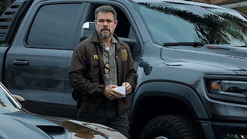 A man in a law enforcement jacket stands outdoors between parked vehicles, holding papers, with a serious expression in a suburban or urban setting.