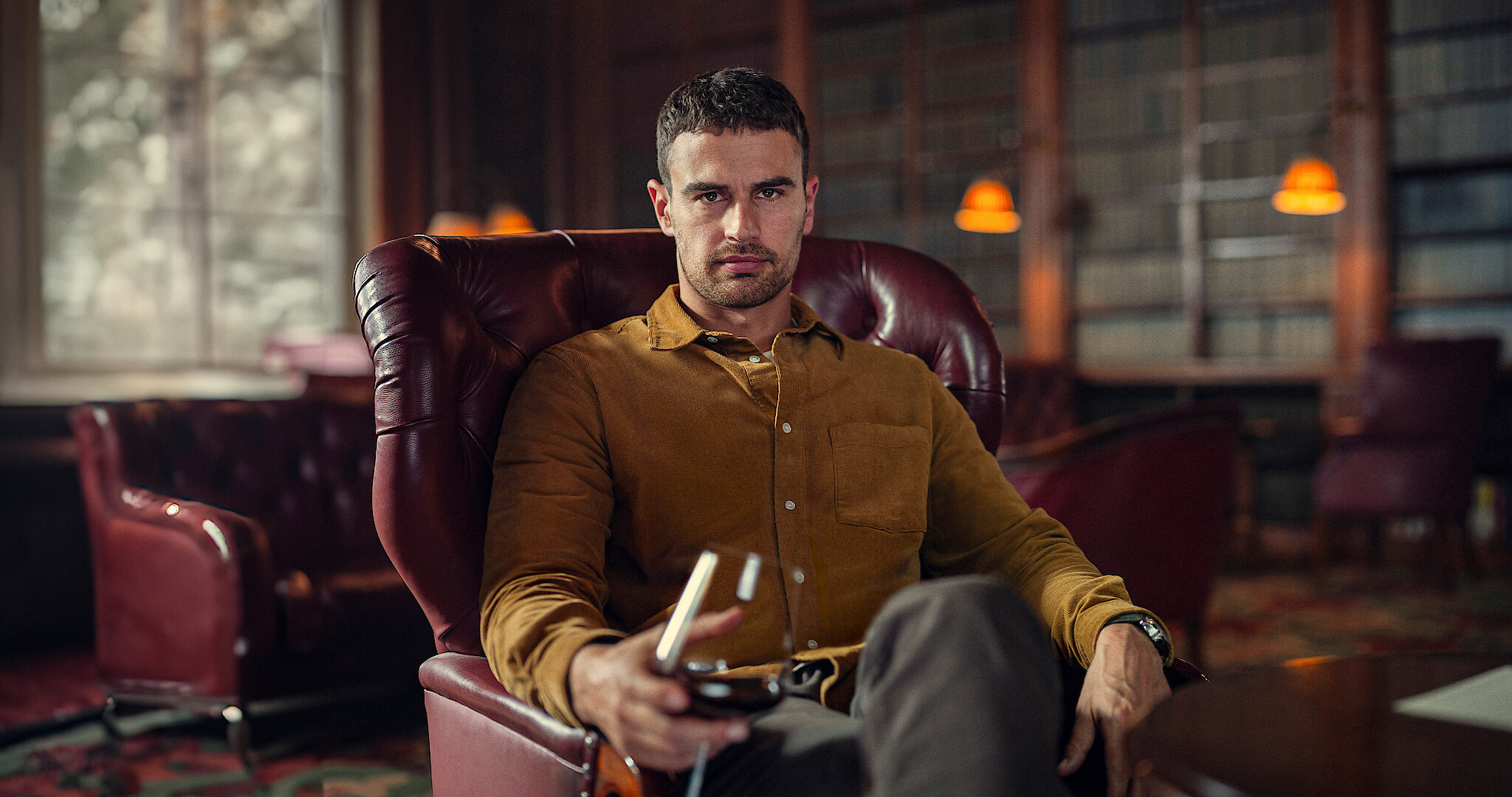 View of a man sitting in a chair, holding a wine glass, staring into the camera confidently. 